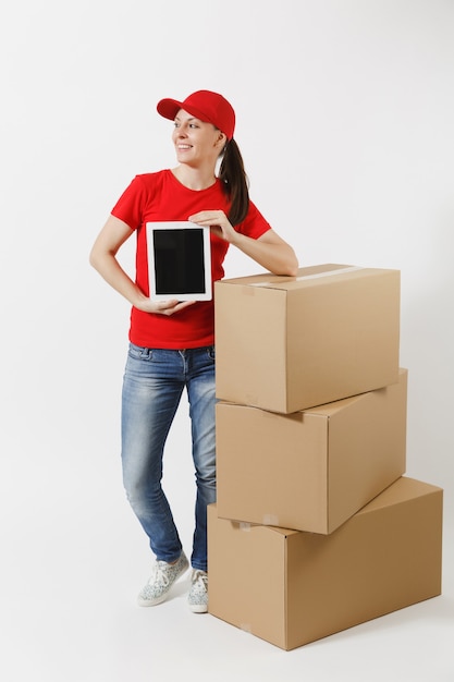 Toute la longueur de la livraison jeune femme au bonnet rouge, t-shirt isolé sur fond blanc. Courrier féminin près de boîtes en carton, ordinateur tablette, écran vide noir vierge. Réception du colis. Espace de copie.