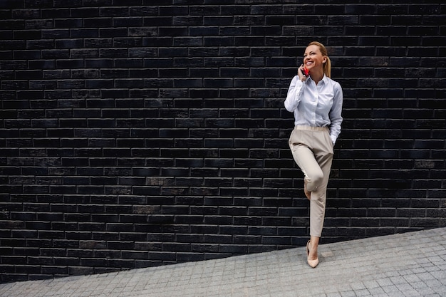 Toute la longueur de la superbe femme d'affaires à la mode blonde appuyée sur le mur et ayant un appel téléphonique.