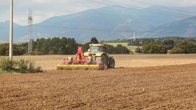 Le tracteur jaune tire le mécanisme rouge de semis au-dessus du champ sec, des arbres et des montagnes à l'arrière-plan