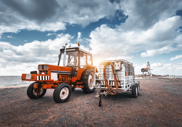 Tracteur rouge avec remorque contre phare et ciel nuageux