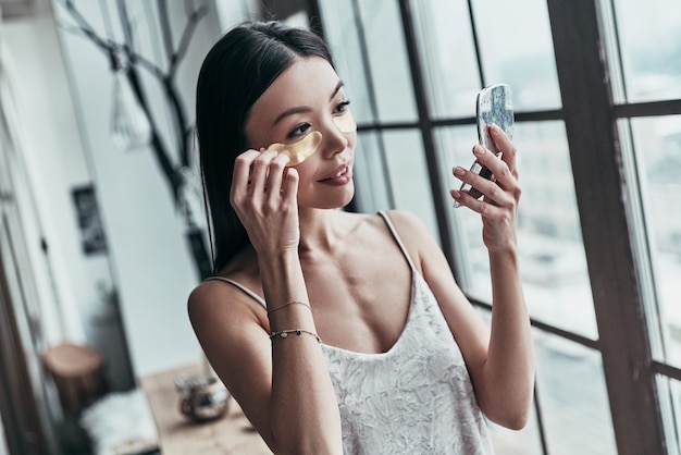 Traitement de beauté. Jolie jeune femme utilisant des cache-œil et souriant tout en passant du temps à la maison