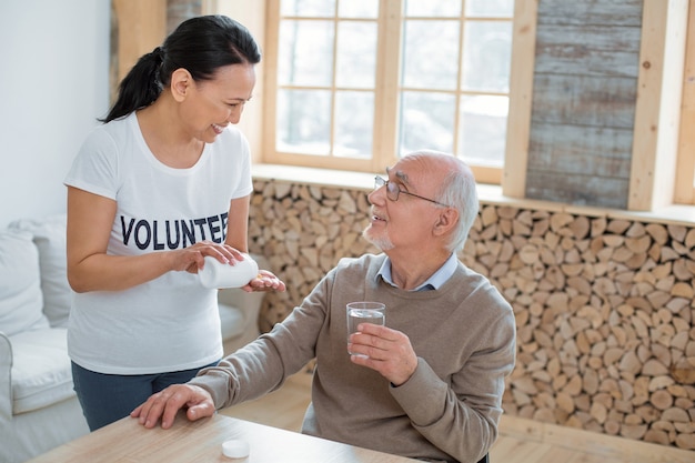 Traitement médical. Joyeux bénévole optimiste debout tout en riant avec un homme senior qui tient un verre d'eau