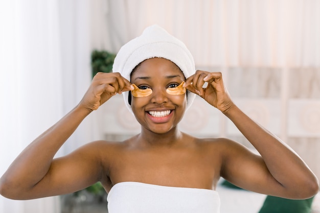 Traitement de la peau des yeux. Jeune femme afro-américaine en serviette blanche sur la tête avec des patchs sous les yeux sur fond clair