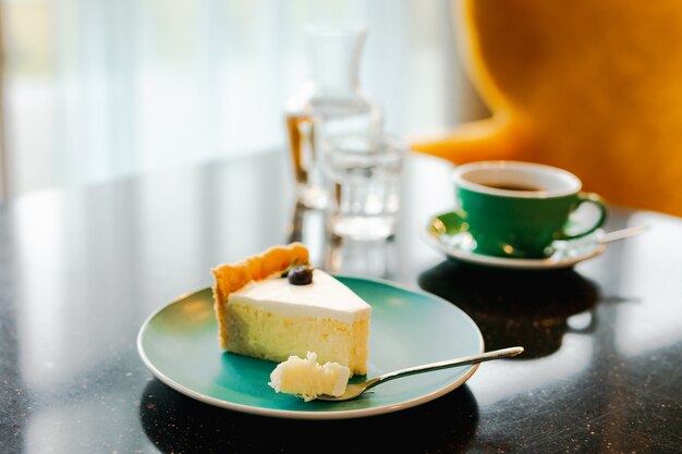 Une tranche de cheesecake sur une assiette bleue, une fourchette à dessert avec un petit morceau de tarte, une tasse de café noir, une carafe d'eau et un verre, sur un tableau noir