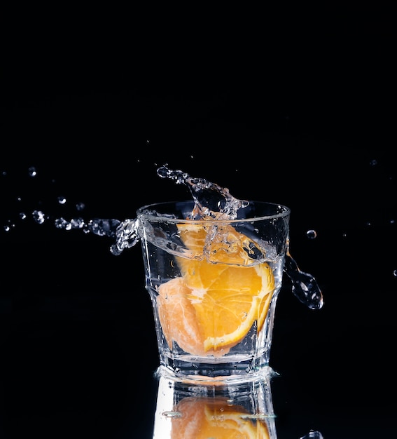 Tranche de citron éclaboussant dans un verre d'eau avec un jet de gouttelettes d'eau en mouvement en suspension dans l'air au-dessus du verre sur un fond sombre.