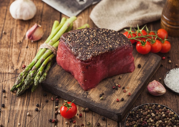 Tranche de dessus de boeuf cru avec sel et poivre sur une planche à découper en bois avec des tomates, de l'ail et des pointes d'asperges sur une table de cuisine en bois