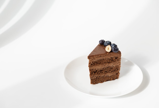 Une tranche de gâteau au chocolat avec des bleuets et des noix sur une plaque blanche sur fond texturé blanc