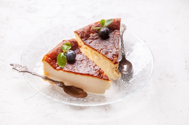 Tranche de gâteau au fromage brûlé basque fait maison sur une assiette avec des myrtilles et des feuilles de menthe sur fond clair.