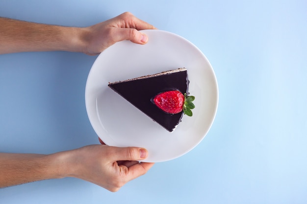 Une tranche de gâteau aux fraises avec glaçage au chocolat dégoulinant dans une assiette blanche en mains sur bleu