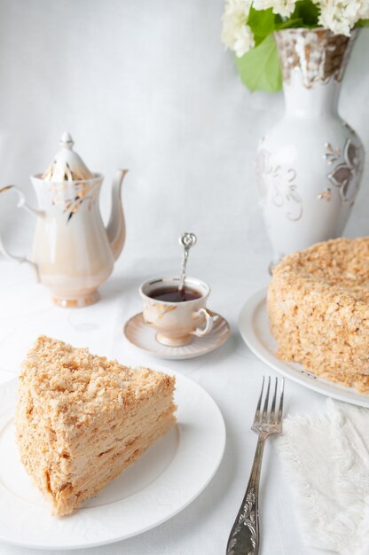 Tranche de gâteau Napoléon multicouche avec crème au beurre Sur une assiette blanche Gros plan À côté d'une assiette se trouve une serviette et une fourchette En arrière-plan se trouve une tasse de théière et un vase de fleurs Fond blanc