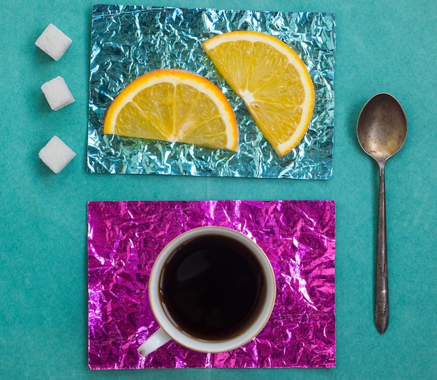 Tranche d'orange et une tasse de café sur des supports en papier d'aluminium jaune et turquoise sur une surface en bois