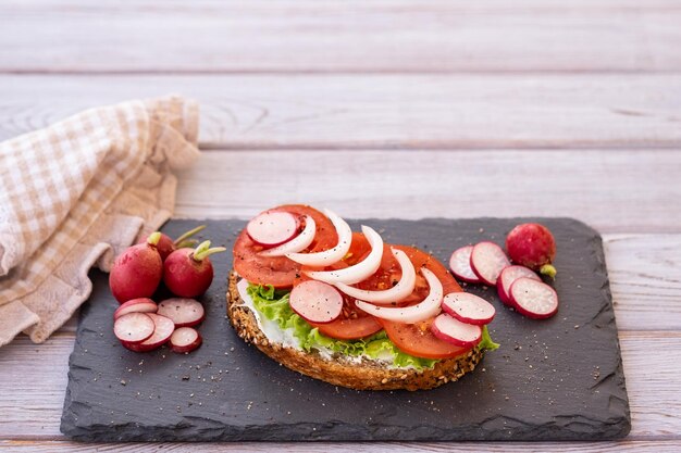 Une tranche de pain aux graines avec du fromage et des tranches de tomates, d'oignons et de radis sur une planche à découper noire. Fond de table en bois