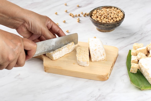 Trancher la main féminine Tempeh ou Tempe sur planche à découper en bois à l'aide d'un couteau. Tempe est un produit de soja fermenté originaire d'Indonésie. Processus de cuisson pour faire du tempeh sauté (Orek)