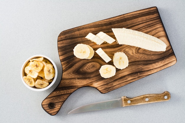 Tranches de banane sur une planche à découper et un couteau et des chips de banane dans un bol sur la table