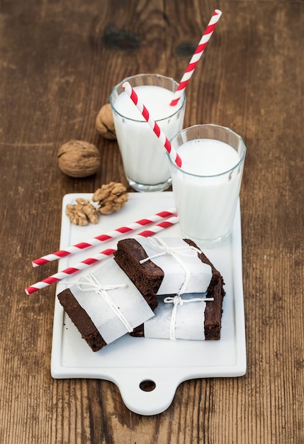 Tranches de brownie au chocolat enveloppées dans du papier et fatiguées avec une corde, des verres de lait, des pailles à rayures, des noix sur un plateau en céramique blanc sur du bois rustique