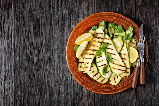 Tranches de courgettes grillées sur une plaque d'argile avec du citron, de l'huile d'olive, des épices et des feuilles de basilic frais sur une table en bois sombre, à plat, espace libre, cuisine italienne