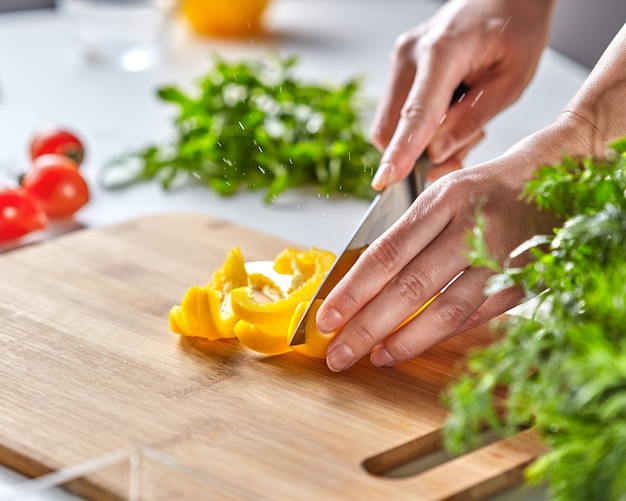 Des tranches juteuses de poivron jaune ont coupé les mains d'une femme en une planche de bois sur une table de cuisine blanche avec des tomates et du persil. Copiez l'espace pour le texte. Cuisiner une salade saine étape par étape