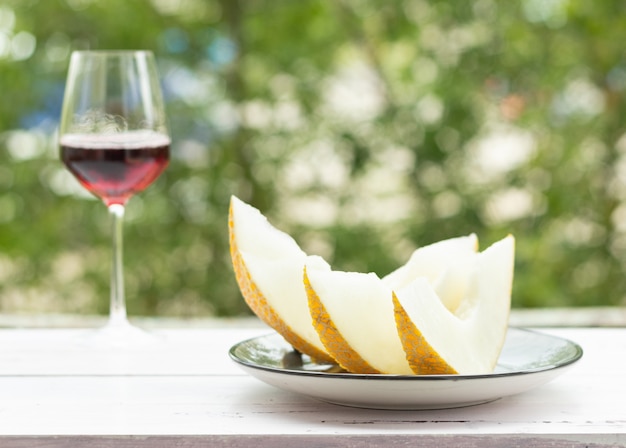 Tranches de melon sur une table en bois blanche, arbres verts sur le fond. Un verre de vin rouge.