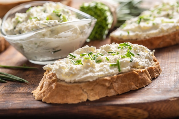 Photo des tranches de pain croustillant et un bol en verre avec une tartinade de fromage à la crème et de la ciboulette coupée sur une planche à découper en bois vintage.