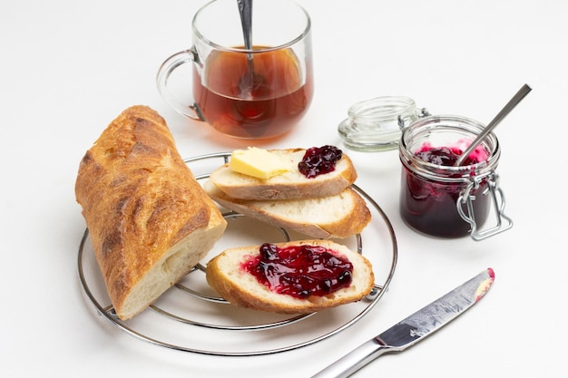 Tranches de pain avec du beurre et de la confiture sur un support en métal. Pot de confiture et verre de thé sur table. Fond blanc. Vue de dessus