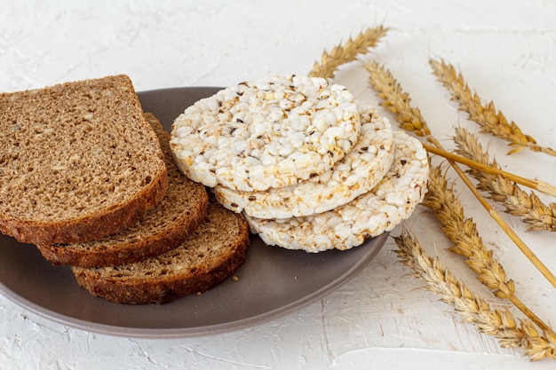 Tranches de pain et galettes de riz soufflé sur l'assiette, épillets de blé sur fond blanc structuré. Vue de dessus.