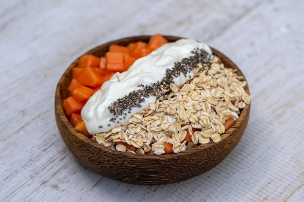 Tranches de papaye sucrée avec flocons d'avoine graines de chia et yaourt blanc dans un bol de noix de coco sur fond de bois blanc gros plan