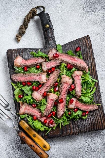 Tranches de steak de boeuf grillé avec salade de feuilles de roquette sur une planche à découper rustique. Fond blanc. Vue de dessus.