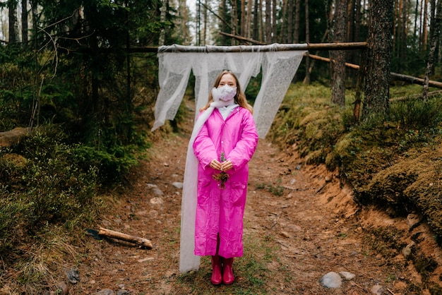 Étrange jeune femme en masque et imperméable rose posant dans la forêt