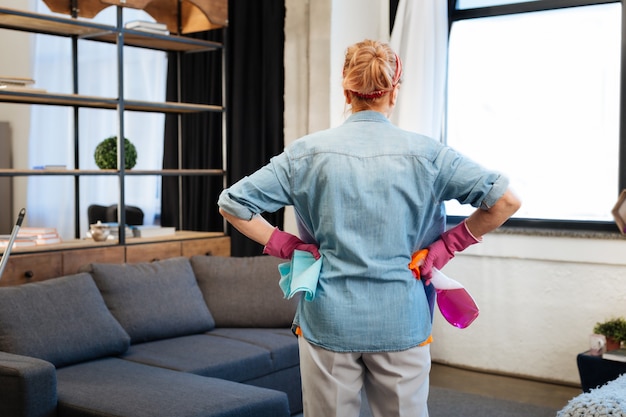 Transporter un spray de nettoyage. Femme résolue aux cheveux attachés observant son appartement et planifiant le processus de nettoyage