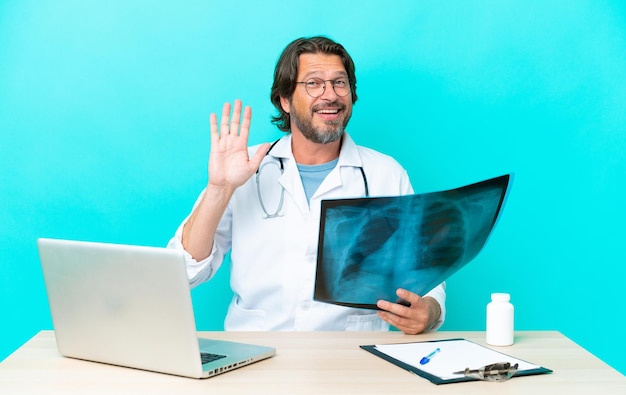 Traumatologue néerlandais senior professionnel au bureau saluant avec la main avec une expression heureuse