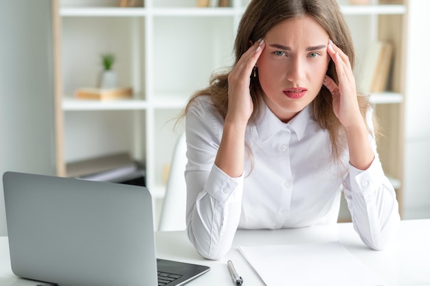 Travail de bureau Femme épuisée Considérer le problème Une dame fatiguée assise au bureau a ouvert un ordinateur portable tenant des temples souffrant de maux de tête à l'intérieur de la pièce lumineuse