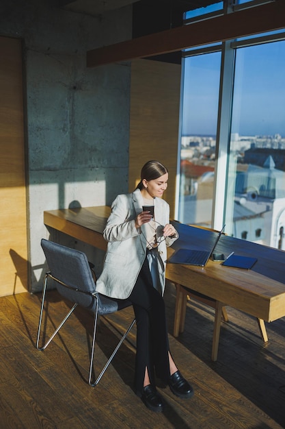 Travail de bureau séduisante jeune femme gestionnaire travaillant au bureau debout dans un espace de travail lumineux à l'aide d'un ordinateur portable moderne jeune femme buvant un délicieux café