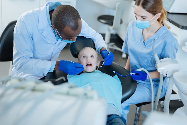 Travail coordonné. Agréable homme dentiste effectuant un examen de sa petite cavité buccale de patients tandis que son infirmière tenant un éjecteur de salive