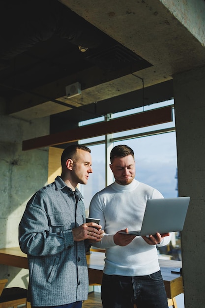 Travail de deux collègues masculins debout ensemble au bureau tout en échangeant des idées avec un ordinateur portable dans la salle de réunion et en analysant les plans de travail Travailler dans un bureau spacieux et moderne