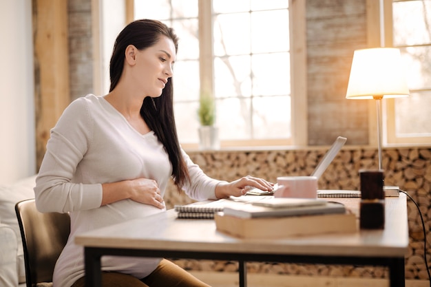 Travail difficile. Charmante femme enceinte sérieuse assise de profil tout en utilisant un ordinateur portable et en mettant la main sur le ventre