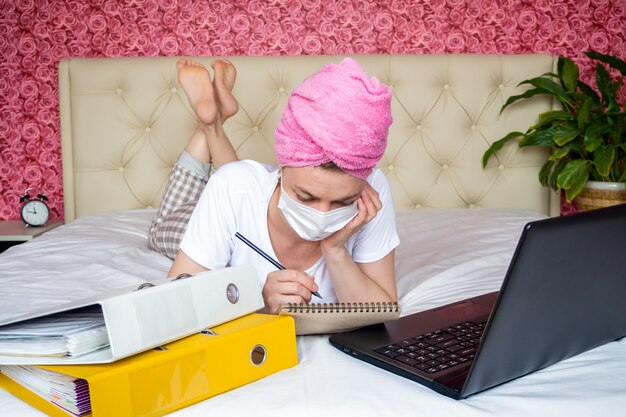 Travail à distance depuis la quarantaine à domicile. Jeune fille de race blanche sur le lit dans un masque à faire leurs devoirs. il regarde son ordinateur portable et prend des notes dans un cahier.