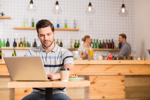 Travail à distance. Smart bel homme positif assis dans le café et manger de la nourriture tout en travaillant sur l'ordinateur portable