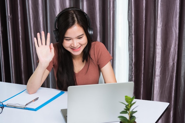 Travail à domicile, jeune femme d'affaires asiatique heureuse souriante assise sur un espace de travail de bureau portant des écouteurs conférence téléphonique vidéo avec ordinateur portable lever la main en disant bonjour à l'équipe au bureau à domicile