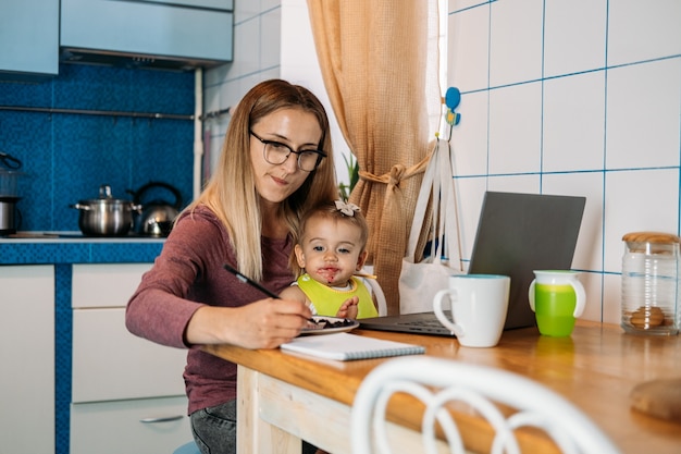 Travail à domicile jeune mère avec petite fille travaillant à la maison à l'aide d'un ordinateur portable sur fond de cuisine jeune