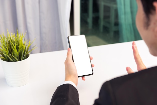 Travail à domicile, vue arrière d'un jeune homme d'affaires asiatique souriant portant un appel de vidéoconférence en costume ou facetime par un écran vierge de téléphone mobile numérique intelligent, levez la main pour dire bonjour à vos coéquipiers sur le bureau