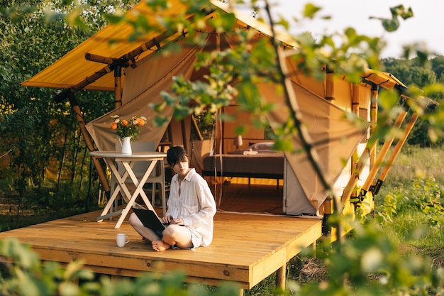 Travail du week-end Une femme commence sa matinée à travailler sur un ordinateur portable tout en étant assise près d'une tente dans un site de glamping pour touristes