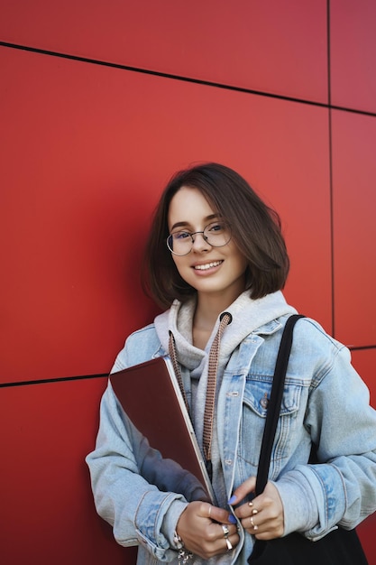 Travail d'éducation et concept de jeunesse Portrait vertical d'une fille queer souriante aux cheveux courts dans des verres regardant la caméra tenant un ordinateur portable près de la poitrine étudiant rubrique bibliothèque préparer l'examen