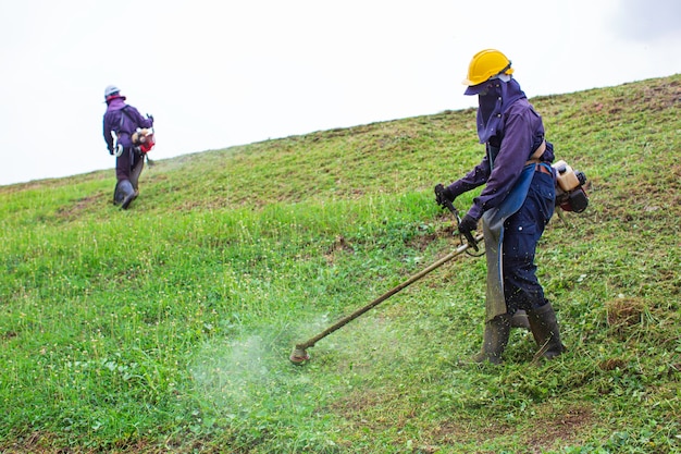 Le Travail Des Femmes Porte Des Vêtements De Protection Tond Sur La Colline L'herbe à Gazon Avec Une Tondeuse à Gazon