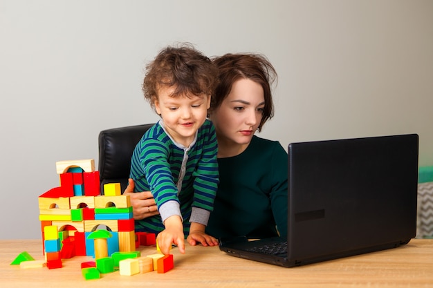 Travail à la maison. Une femme est assise à travailler sur un ordinateur portable pendant qu'un enfant joue à des cubes et construit une grande maison à plusieurs étages.