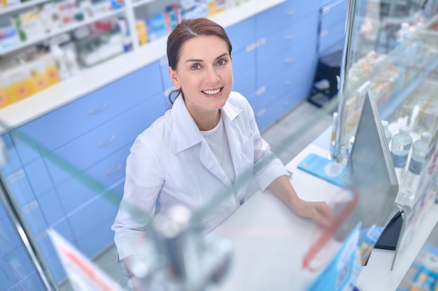 Travail, positif. Souriante jolie jeune femme adulte en blouse blanche debout près de la caisse enregistreuse de la pharmacie en levant