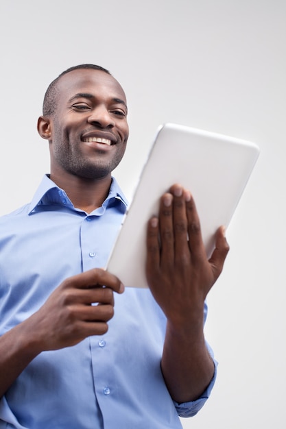Travail sur tablette numérique. Vue à faible angle d'un bel homme africain travaillant sur une tablette numérique et souriant tout en se tenant isolé sur fond gris