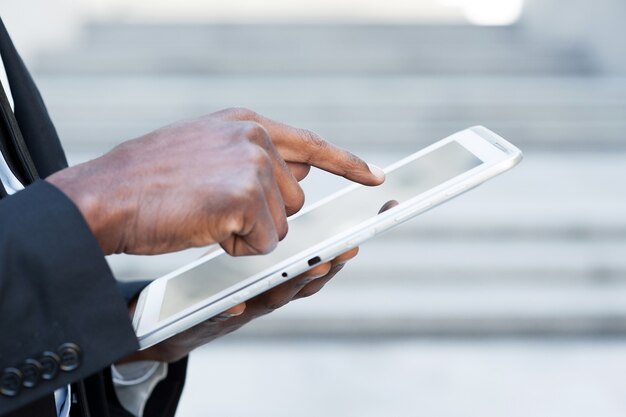 Travail sur tablette. Vue latérale image recadrée d'un homme africain en tenue de soirée travaillant sur une tablette numérique tout en se tenant à l'extérieur