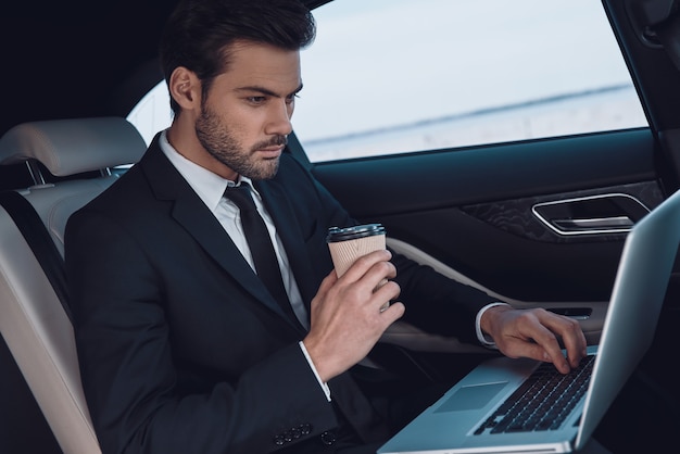 Travail en voiture. Beau jeune homme en costume complet travaillant à l'aide d'un ordinateur portable alors qu'il était assis dans la voiture