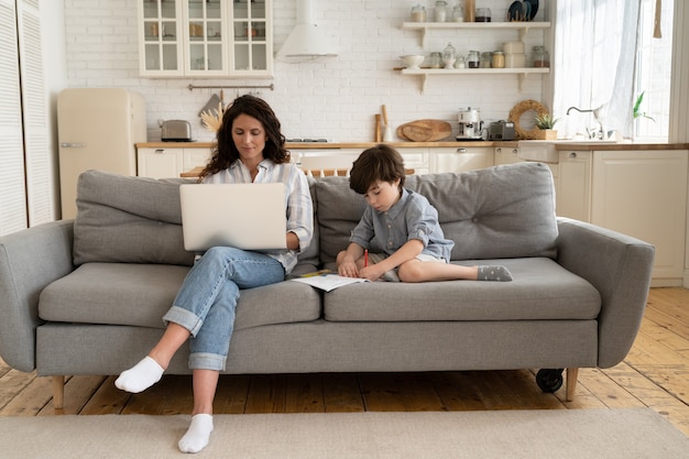 Travaillant à domicile, une mère indépendante utilise un ordinateur portable assis sur un canapé avec un petit fils dessinant