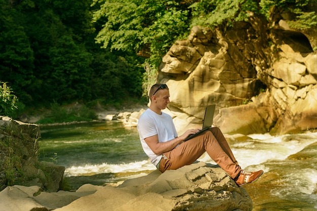 Travailler dans le concept de la nature. Homme avec ordinateur portable assis au bord de la rivière sur la cascade et les arbres verts. Vue de côté